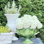 A green ceramic artichoke-shaped planter filled with white hydrangea blooms, displayed on a dark gray slatted table outdoors with other white planters and green foliage in the background.