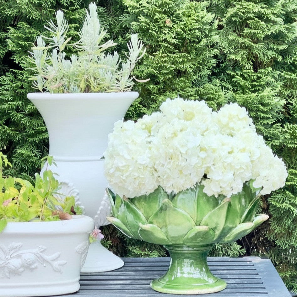 A green ceramic artichoke-shaped planter filled with white hydrangea blooms, displayed on a dark gray slatted table outdoors with other white planters and green foliage in the background.