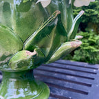 A glossy green ceramic flower vase shaped like an artichoke holding white flowers, set on a dark slatted table with blurred green foliage in the background.