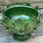 A green glazed ceramic artichoke-shaped  bowl with a pedestal base, photographed outdoors on stone pavement against a stone wall.