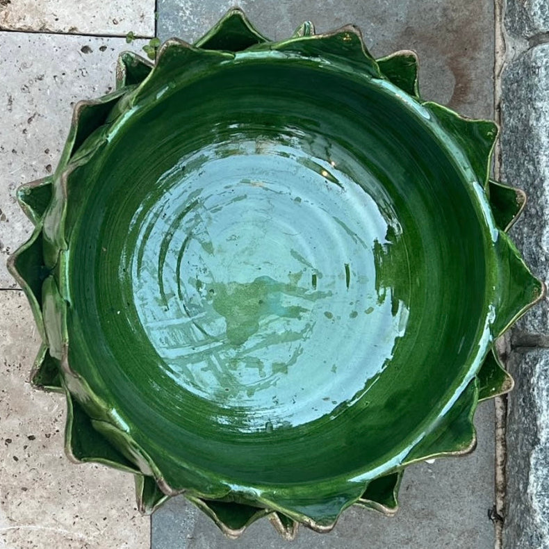 A handmade ceramic bowl with a scalloped, lotus-like rim, glazed in a glossy dark green glaze, photographed from directly above on a stone tile surface.