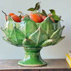 A green glazed ceramic pedestal bowl shaped like an artichoke, filled with oranges and green leaves, displayed on a wooden surface against a light gray wall.