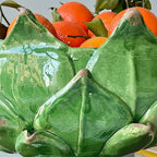 A close-up of a glossy green ceramic lotus-shaped bowl filled with bright orange citrus fruits and green leaves.