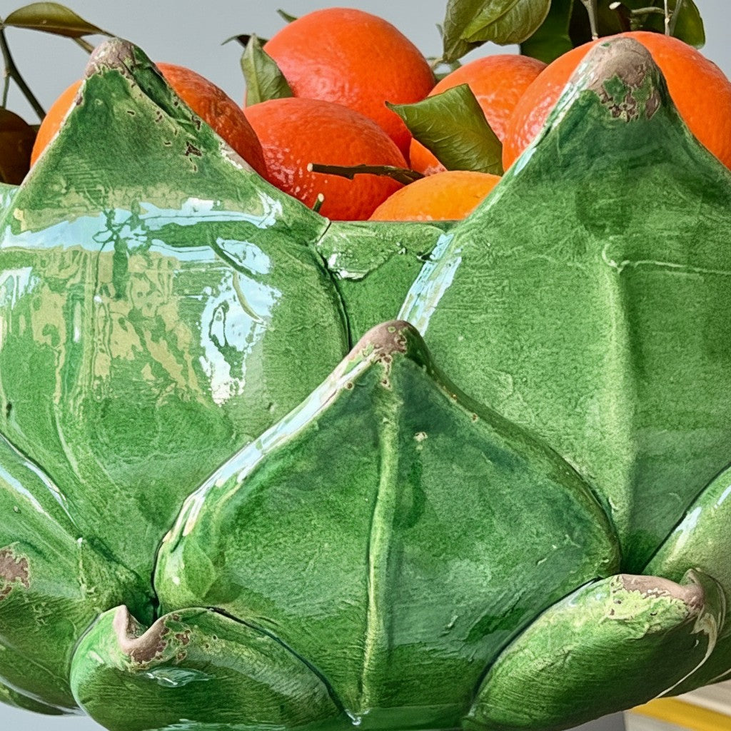 A close-up of a glossy green ceramic lotus-shaped bowl filled with bright orange citrus fruits and green leaves.
