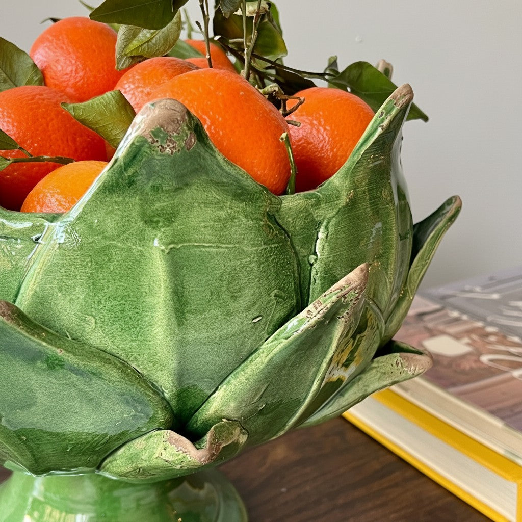 A green ceramic bowl shaped like a lotus flower, filled with bright oranges and green leaves, set on a wooden surface with books in the background.