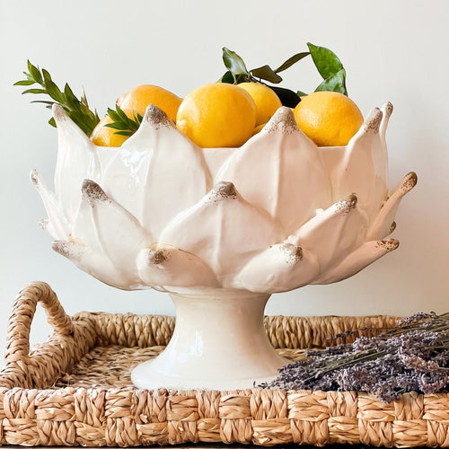 Heirbloom Studio off-white ceramic artichoke pedestal bowl filled with fresh lemons on a woven tray.