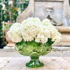Heirbloom Studio handcrafted emerald green majolica artichoke pedestal bowl filled with white hydrangeas outdoors.