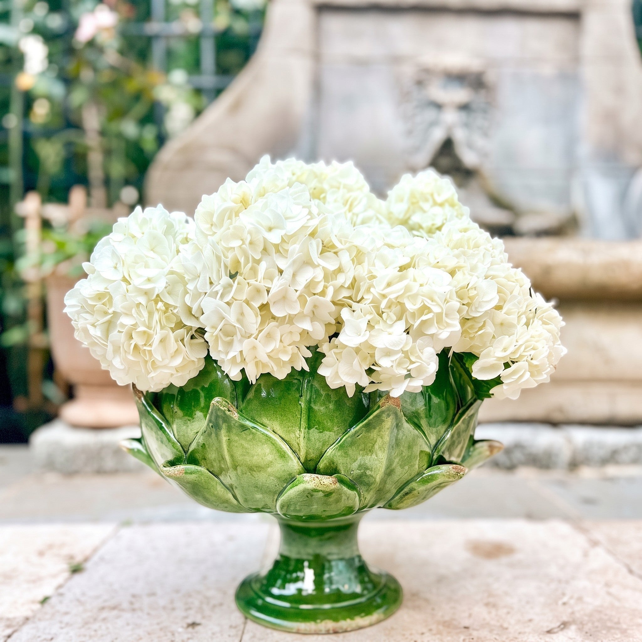 Heirbloom Studio handcrafted emerald green majolica artichoke pedestal bowl filled with white hydrangeas outdoors.